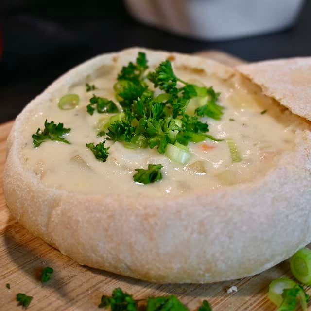 Eine rustikale Schüssel aus einem ausgehöhlten Brotlaib, gefüllt mit cremiger Kartoffelsuppe und mit frischen Kräutern dekoriert.