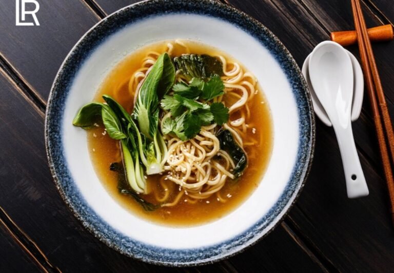 Ein Teller mit dampfender Ramen-Nudelsuppe, garniert mit frischem Pak Choi, Koriander und Sesam, neben Essstäbchen und einem Löffel auf einem dunklen Holztisch.